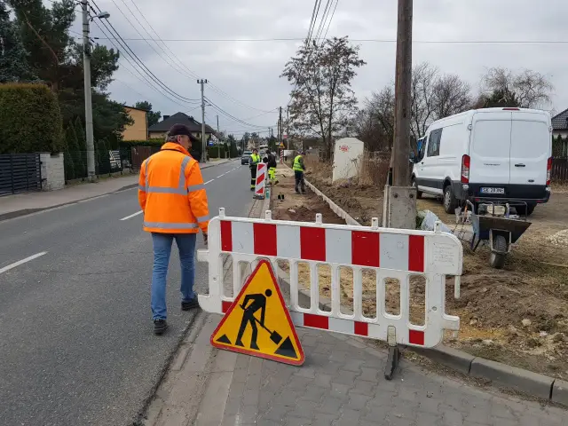 Ekipa remontowa pracuje przy drodze, ogrodzona barierami i znakiem ostrzegawczym.