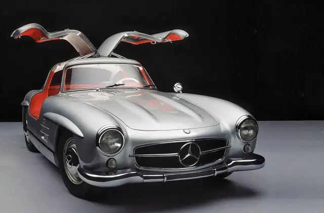 Srebrny Mercedes 300 SL z otwieranymi do góry drzwiami, z czerwonym wnętrzem, prezentuje się elegancko na ciemnym tle.