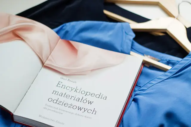 Książka "Encyklopedia materiałów odzieżowych" obok błękitnej i granatowej bluzki na wieszakach. Dowiedz się, jak czytać etykiety ubrań, żeby rozpoznać poliester od wiskozy.