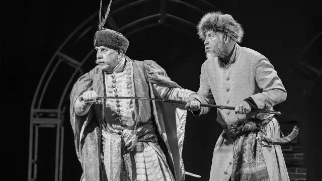 Dwóch mężczyzn w historycznych strojach, jeden z szablą, drugi z fuzją. Ich miny sugerują komizm w zemście, gdy przygotowują się do konfrontacji.