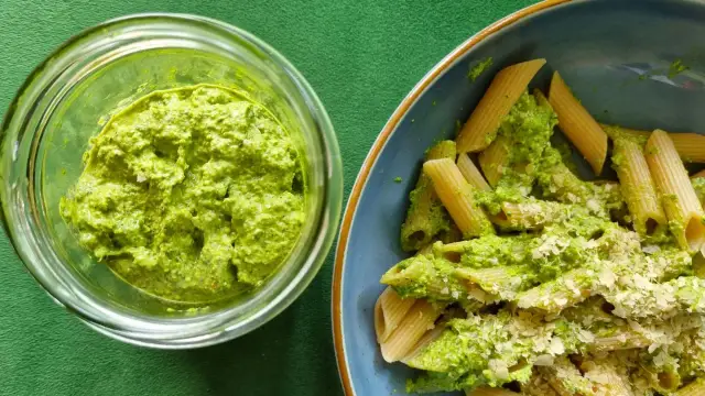 Pesto z liści rzodkiewki w słoiku obok talerza penne z tym zielonym sosem i startym parmezanem.