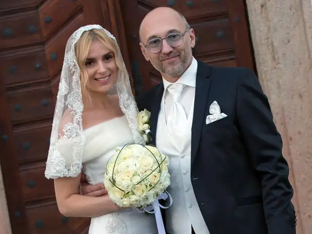 Szczęśliwa panna młoda z bukietem i jej mąż, w eleganckich strojach ślubnych, pozują przed drewnianymi drzwiami.