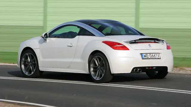 Biały Peugeot RCZ z czarnym dachem pędzi po drodze, za zielonym ekranem dźwiękochłonnym.