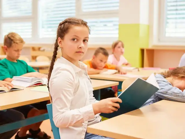 Dziewczynka z warkoczem czyta książkę, podczas gdy inni uczniowie siedzą przy ławkach. To część programu nauczania edukacja zdrowotna.