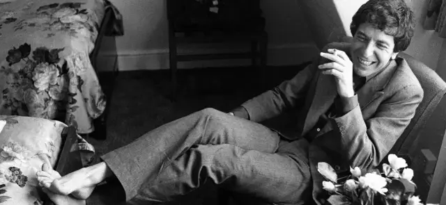 Uśmiechnięty Leonard Cohen, palący papierosa, w garniturze. W tle kwieciste tkaniny, jakby inspirowane jego ponadczasowymi utworami.
