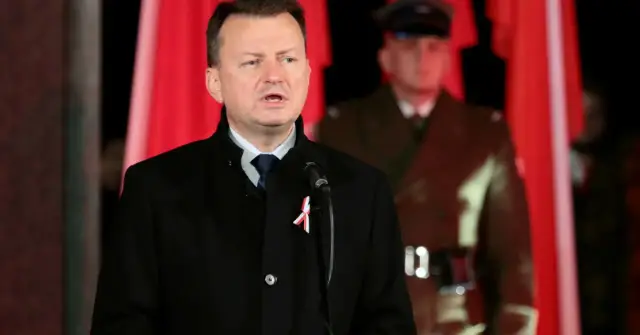 Mężczyzna w czarnym płaszczu z biało-czerwoną wstążką przemawia przy mikrofonie. W tle widać żołnierza i polskie flagi. Nie wiadomo, ile lat ma błaszczak.