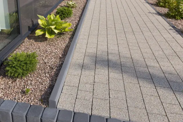 Nowy chodnik z kostki brukowej i obrzeży betonowych, otoczony kamyczkami i zielenią. Widać, ile betonu pod obrzeże zostało użyte do stworzenia solidnej podstawy.