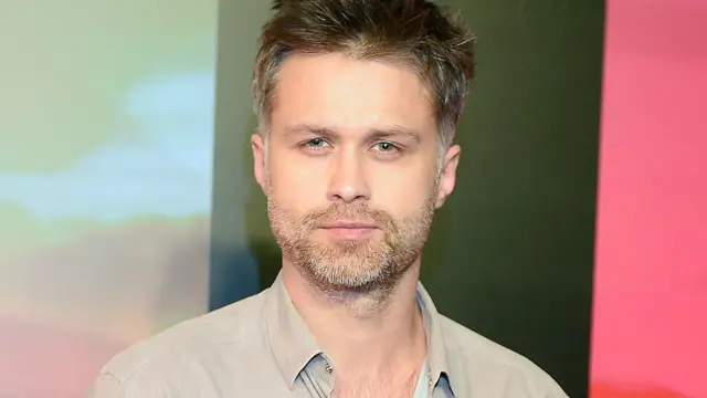 Maciej Zakościelny, aktor, z zarostem i w jasnej koszuli, na tle kolorowych paneli.