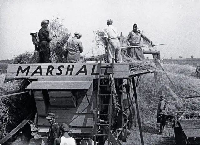 Ludzie pracują przy młocarni "MARSHALL", zbierając plony. To obraz ciężkiej pracy, jakiej wymagał plan Marshalla.