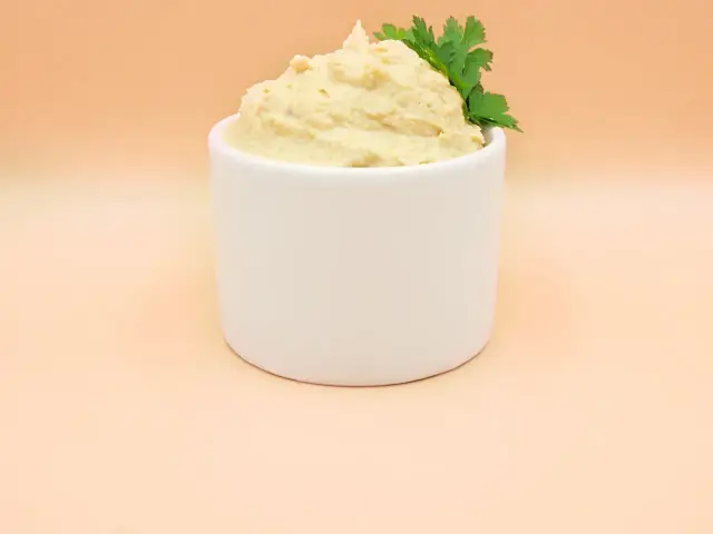 Puszysty hummus z ciecierzycy, udekorowany gałązką natki pietruszki, w białej miseczce.
