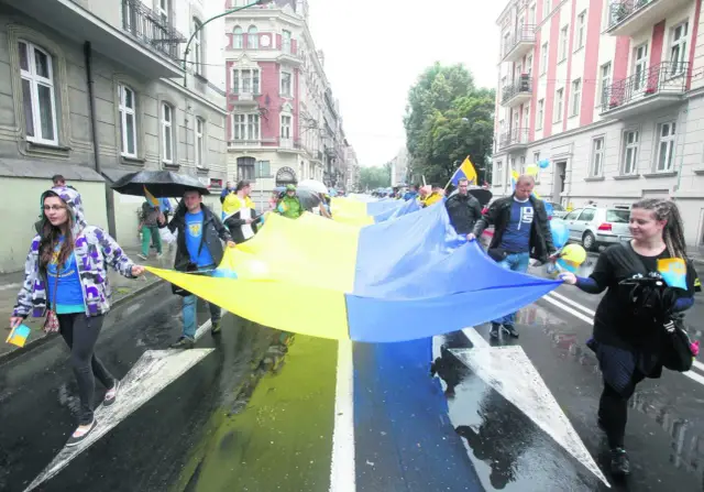 Ludzie niosą wielką flagę w barwach Ukrainy. W deszczu idzie grupa, wśród nich młoda kobieta, która nie jest gorolem.