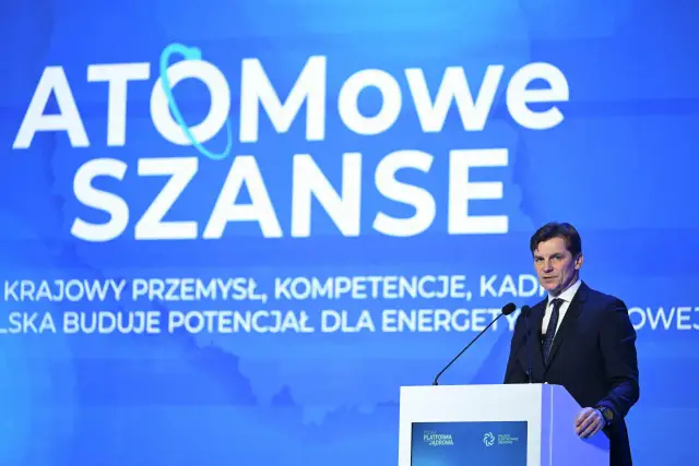 Mężczyzna w garniturze przemawia na tle napisu "ATOMOWE SZANSE", podkreślając rozwój polskiego przemysłu i kompetencji dla energetyki jądrowej.