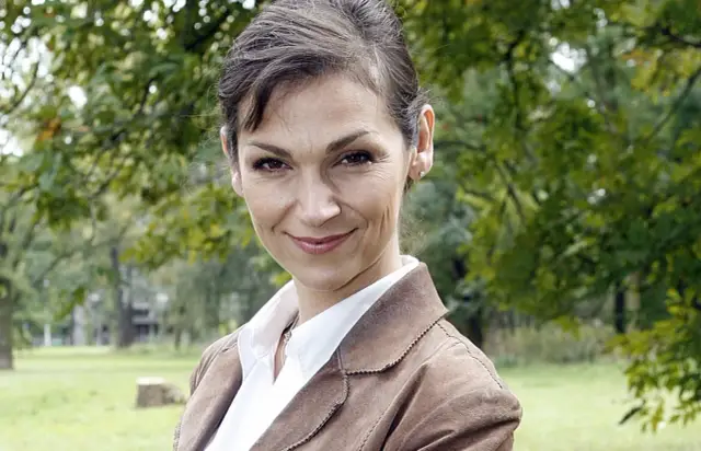 Olga Bończyk w brązowej marynarce i białej koszuli, uśmiecha się na tle drzew.