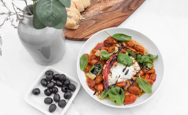Pyszna burrata z pieczonymi pomidorkami koktajlowymi, czosnkiem i bazylią, skropiona octem balsamicznym. Obok czarne oliwki i kromki chleba.