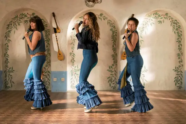 Trzy kobiety w dzwonach z falbanami śpiewają na scenie, jakby szykowały się na Mamma Mia 3.