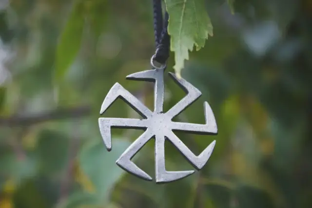 Metalowy symbol, przypominający swastykę słowiańską, wisi na tle zielonych liści.