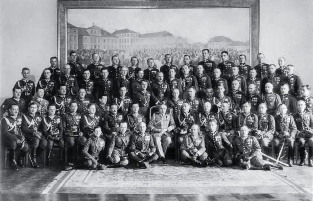 Grupa mężczyzn w mundurach wojskowych, być może z obozu zwolenników Piłsudskiego, pozuje do zdjęcia na tle obrazu.