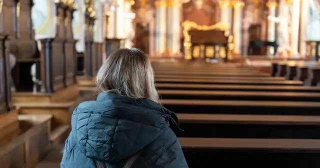 Kobieta w kościele, siedząc w ławce, zastanawia się, co to jest modlitwa. Wnętrze świątyni, pełne ozdób, sprzyja zadumie.