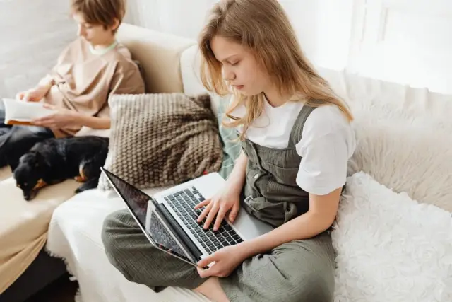 Dziewczynka na laptopie szuka informacji, jak sprawdzić lokalizację dziecka. Na kanapie obok pies i chłopiec z książką.