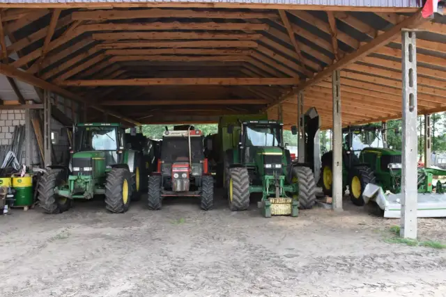 Drewniana wiata bez pozwolenia dla rolnika chroni cztery traktory, w tym zielone John Deere i czerwony Zetor, gotowe do pracy na polu.