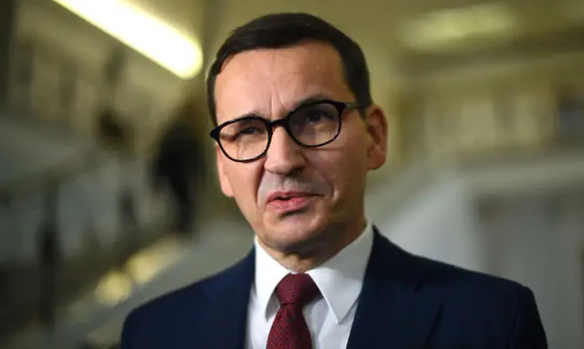 Mateusz Morawiecki w okularach i garniturze, w tle rozmyte korytarze. Dyskusja o majątku Morawieckiego trwa.