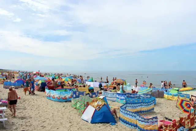 Słoneczna pogoda na sierpień nad morzem. Tłumy plażowiczów, kolorowe parawany i namioty chronią przed wiatrem.