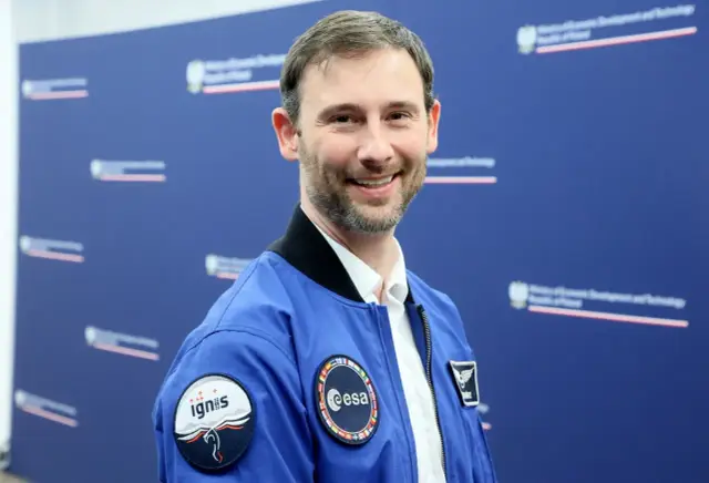 Uśmiechnięty mężczyzna w niebieskiej kurtce z naszywkami ESA i Ignis, jak prawdziwi astronauci, pozuje na tle niebieskiej ściany z logo ministerstwa.