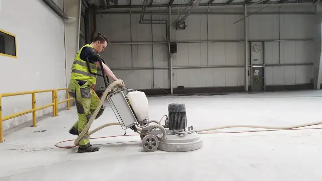 Pracownik w kamizelce odblaskowej szlifuje beton maszyną. To pokazuje, czym szlifować beton, by uzyskać gładką powierzchnię.