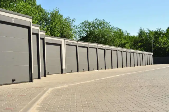 Rząd nowoczesnych, szarych garaży betonowych prefabrykowanych, każdy z numerem, stoi wzdłuż utwardzonego podjazdu, otoczony zielenią.