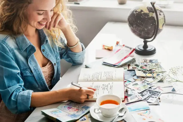 Kobieta pisze w dzienniku, planując podróż. Na stole globus, zdjęcia, bilety i herbata. To inspiracja, jak napisać esej po angielsku o swoich marzeniach.