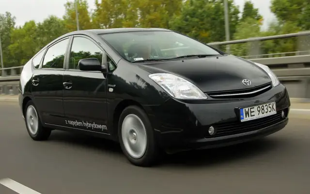 Czarny Toyota Prius 2 z napędem hybrydowym jedzie po drodze, otoczony zielenią.