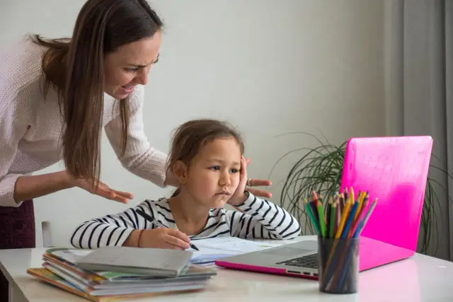 Zestresowana dziewczynka z zeszytem i laptopem, podczas gdy mama z niezadowoloną miną tłumaczy jej coś o ocenach w szkole.