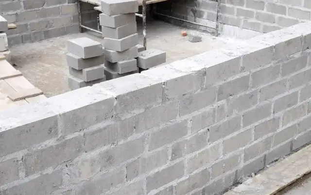 Ściana z bloczków fundamentowych w trakcie budowy. Widać stosy bloczków, gotowe fragmenty muru i materiały budowlane.