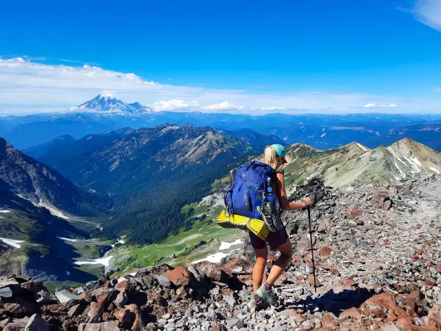 Kobieta z plecakiem wspina się po kamienistym szlaku, podziwiając widok na góry i ośnieżony wulkan. To fragment Pacific Crest Trail.