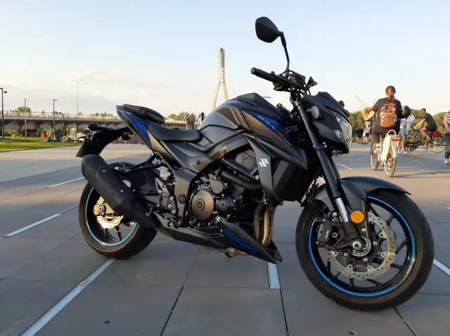 Czarny motocykl Suzuki GSX 750 na tle mostu. W tle ludzie spacerują i jeżdżą na rowerach.
