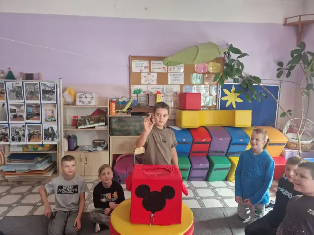 Chłopiec trzyma żołądź, symbol jesieni, przed pudełkiem z Myszką Miki. Dzieci patrzą z zaciekawieniem.