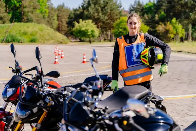 Dziewczyna w kamizelce z literą "L" i kaskiem w ręku gotowa do jazdy na motocyklu. Idealny motocykl na egzamin kat A czeka!
