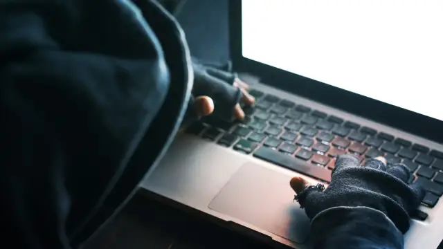 Osoba w kapturze i rękawiczkach pisze na laptopie. To może być początek kradzieży tożsamości.