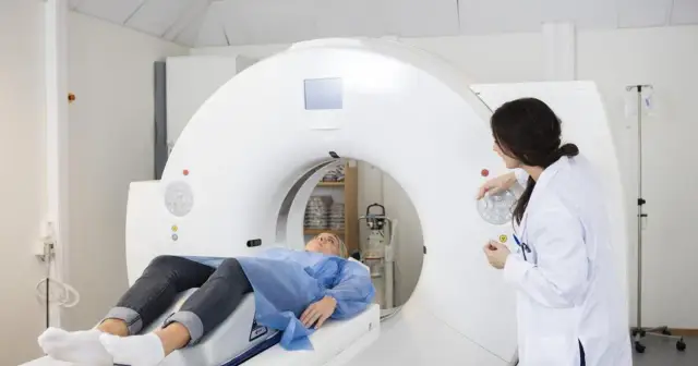 Pacjentka leży na stole tomografu, przygotowana do badania angio-tk tętnic wieńcowych. Lekarka obsługuje aparat.