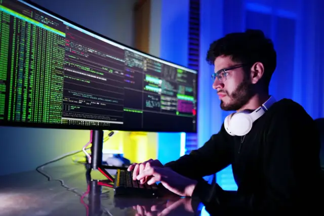 Młody technik programista w okularach, z słuchawkami na szyi, pochyla się nad monitorem z kodem.