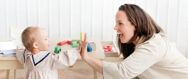 Uśmiechnięta opiekunka daje piątkę dziecku przy stoliku z zabawkami. Idealny stosunek, ile dzieci przypada na jednego opiekuna w żłobku.