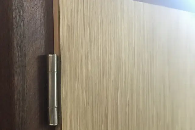 Nowe nakładki na stare futryny drewniane, imitujące jasne drewno, z widocznym metalowym zawiasem.