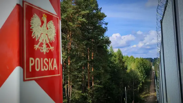 Słup graniczny z orłem i napisem Polska przy lesie. Dalej widać ogrodzenie z drutu kolczastego, symbolizujące strefę **dmz**.