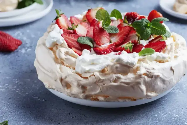 Pyszny tort bezowy z mascarpone i bitą śmietaną, ozdobiony świeżymi truskawkami i miętą.