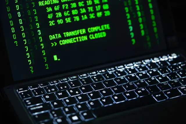 Zielony tekst na ekranie laptopa, "DATA TRANSFER COMPLETE >> CONNECTION CLOSED", sugeruje zakończenie operacji, być może związanej z szyfrowaniem BitLocker.
