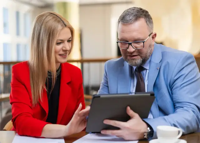 Kobieta w czerwonej marynarce i mężczyzna w niebieskiej analizują dane na tablecie, omawiając strategię biznesową. Ich wspólna praca nad projektem MBA przynosi efekty.