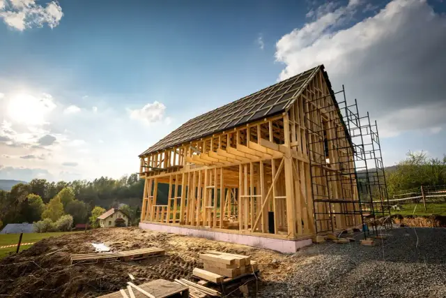 Budowa domu szkieletowego w malowniczej okolicy. Drewniana konstrukcja piętrowego budynku z dachem pokrytym papą, otoczona rusztowaniami.