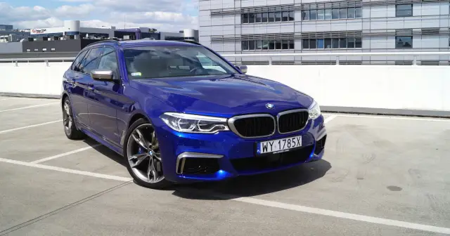 Błękitne BMW 550d kombi na parkingu przed nowoczesnym budynkiem.