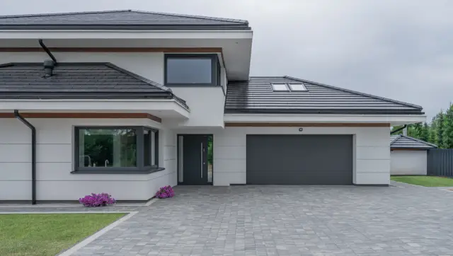 Nowoczesny dom z szarym dachem i drzwiami garażowymi. Duże okna zapewniają światło, a gwarancja na okna daje spokój.