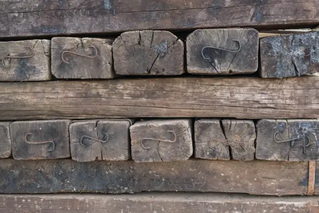 Stare, drewniane podkłady kolejowe w ogrodzie, tworzące rustykalną ścianę. Widać metalowe zaczepy.